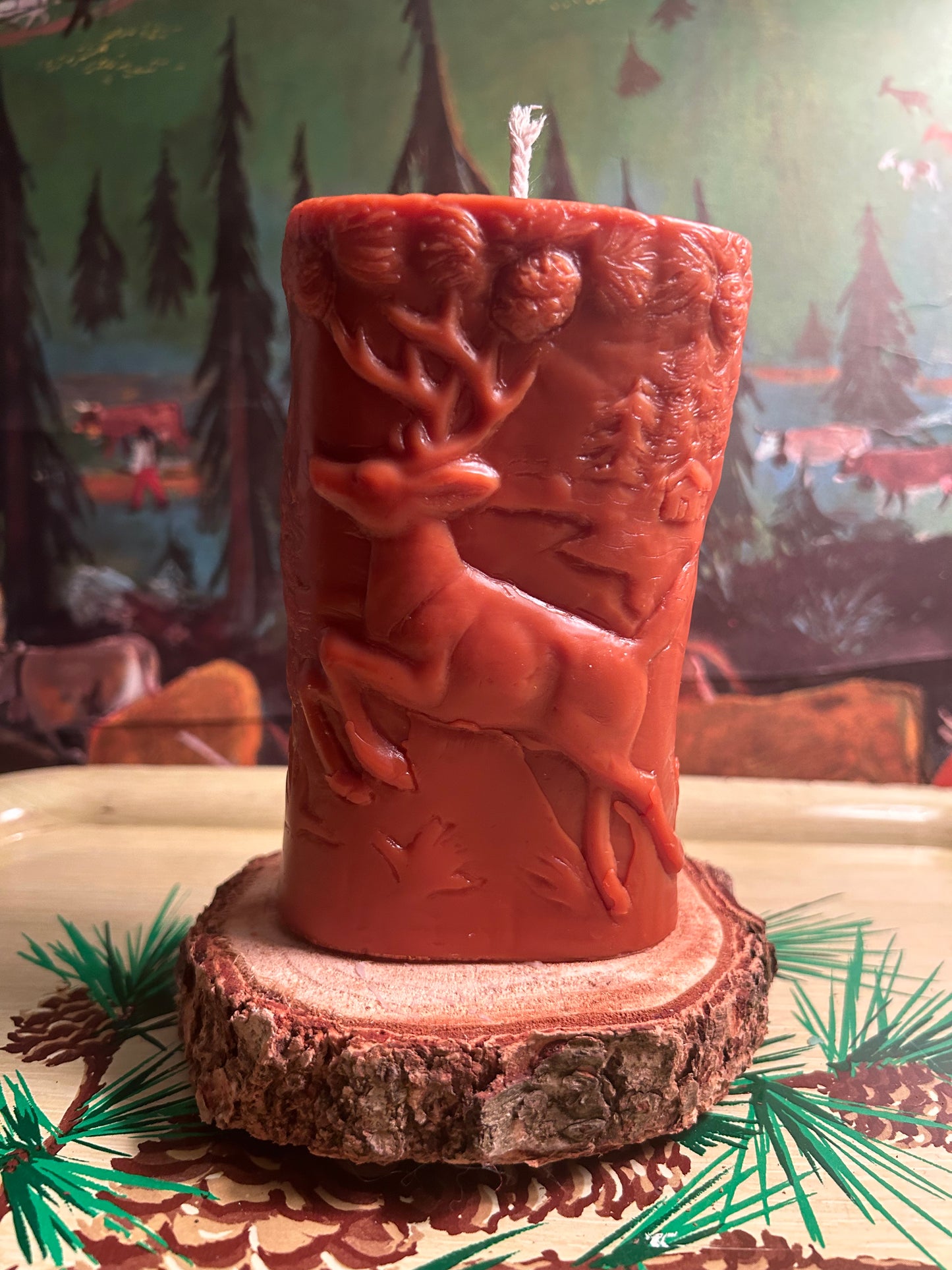 A dark brown molded candle with a stag on one side and a bear on the other side and decorated with pinecones and trees.  Approximately 61/2 inches tall.  Pine trees in the background.