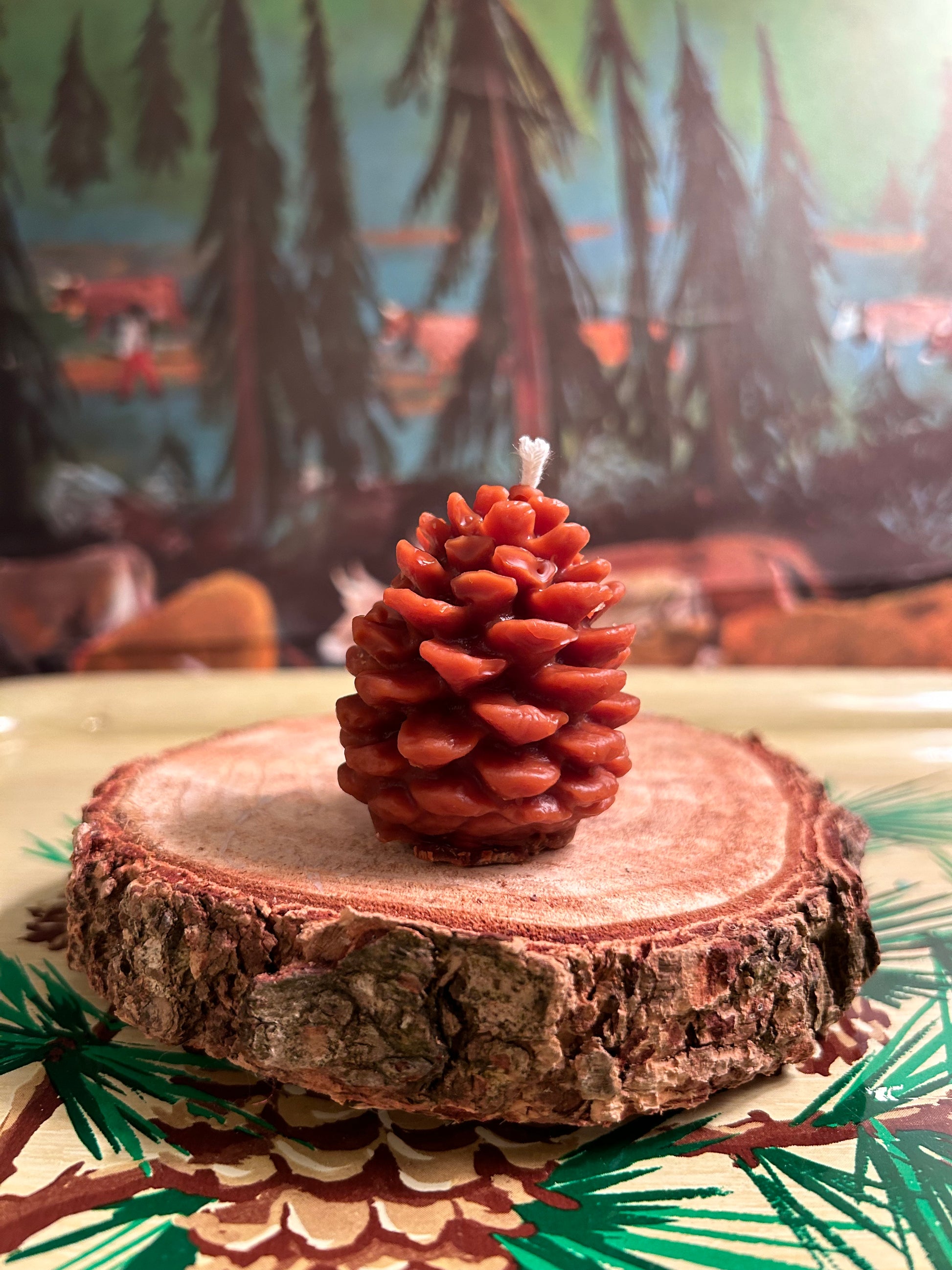 A tiny brown beeswax pinecone molded candle two inches tall sitting on a piece of wood with pine trees in the background.