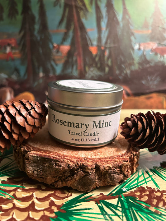 Rosemary Mint travel candle in a 4 oz. tin container sitting on a piece of wood with pinecones and pine trees in the background.