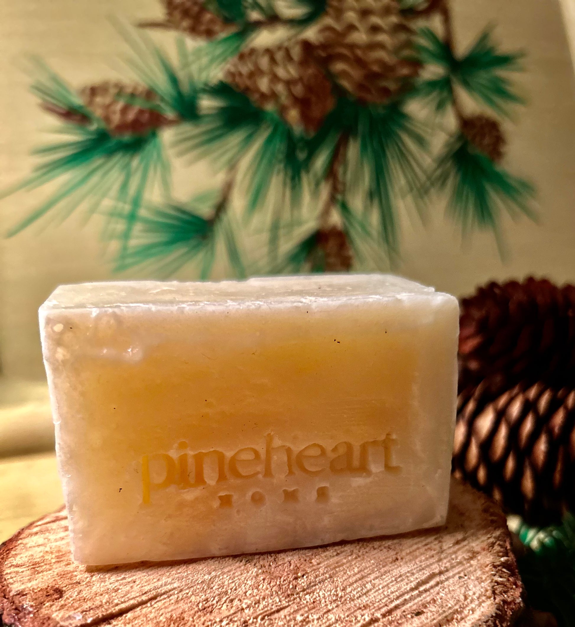 A square white bar of soap sitting on a piece of wood with pinecones in the background.